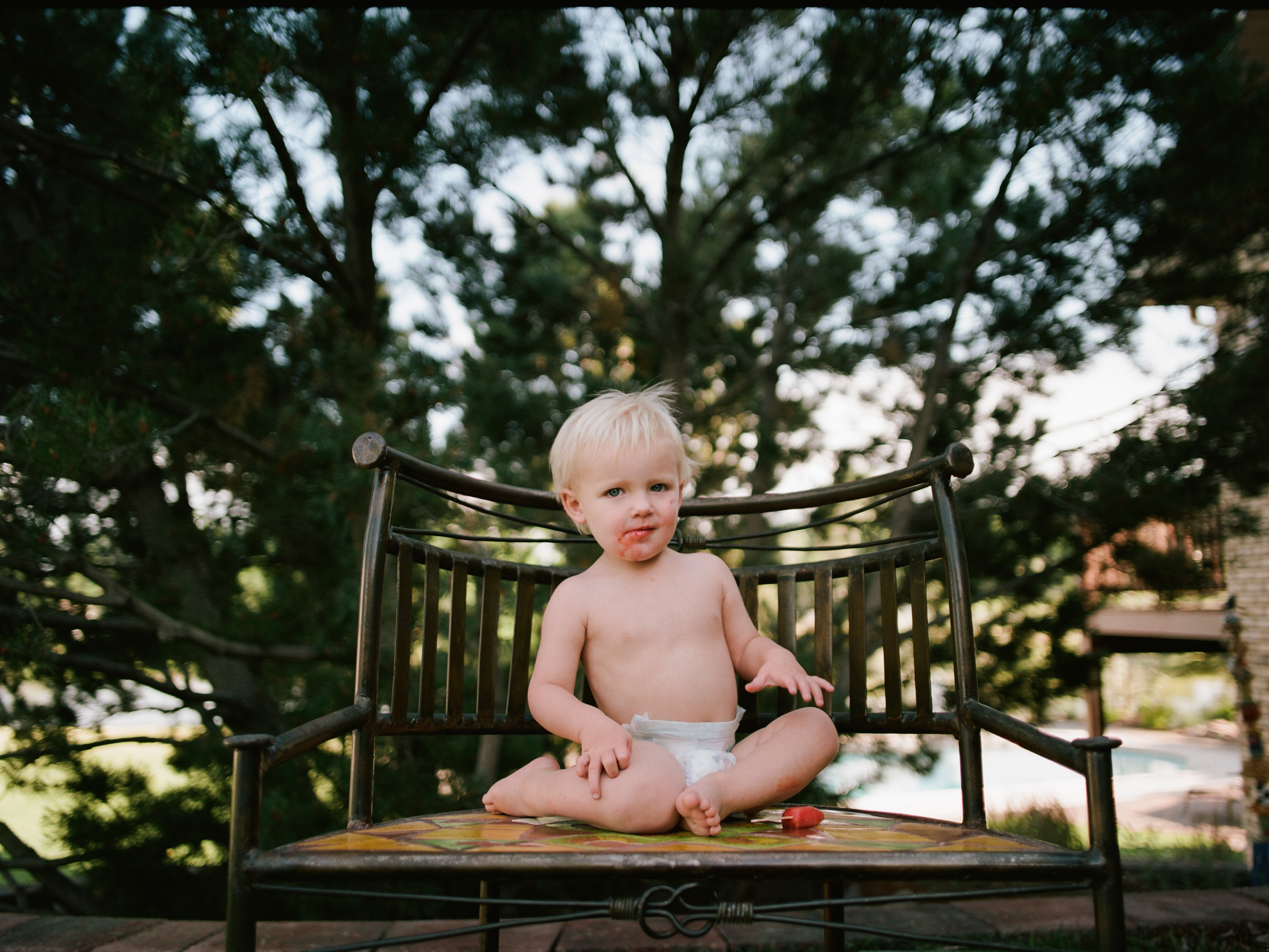 A toddler in diapers sits on a metal chair and eats a popsicle