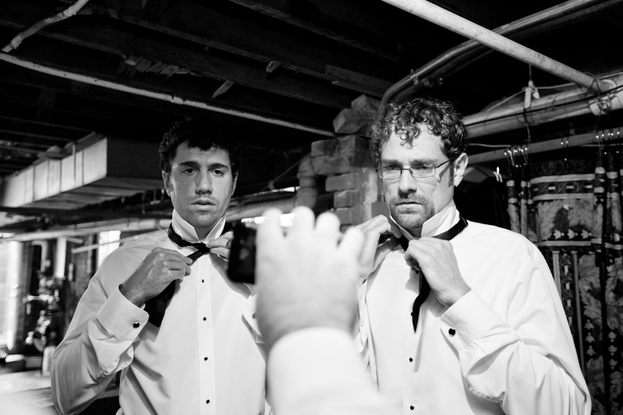 a groom and a groomsman look at a phone while tying bowties