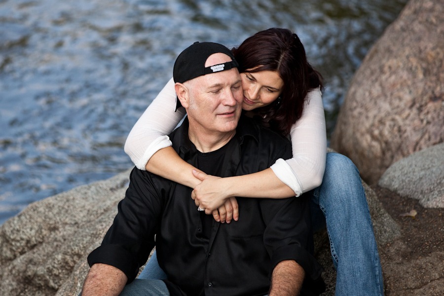 a woman sitting on a rock wraps her arms around a man with a backwards ball cap on