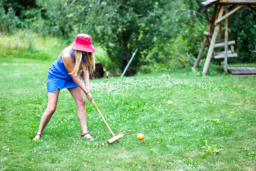 woman in blue dress and red hats hits a yellow ball with a croquet stick