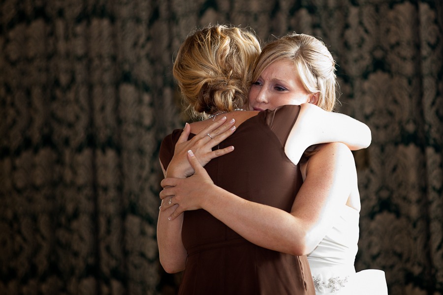 a bride hugs a bridesmaid in a brown dress in front of heavy curtains