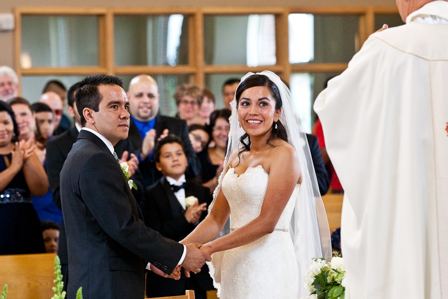 bride and groom hold hands in front of a priest in a church