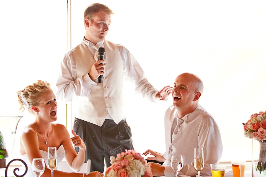 a man in a white vest holds a mic and makes a seated bride and groom laugh