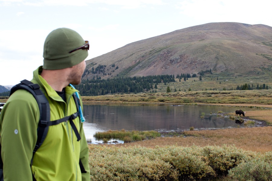 man in green hat and coat looks at a moose standing on the edge of a pond in the mountains