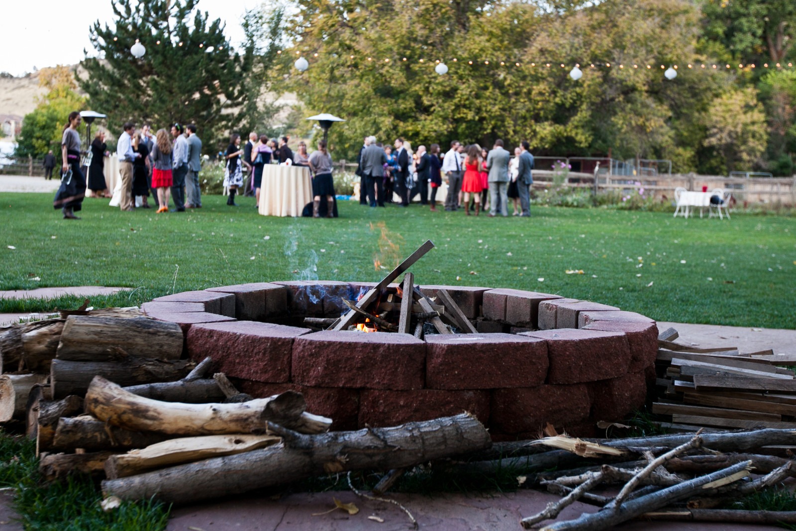 a fire burns in a brick fire pit while wedding guests mingle