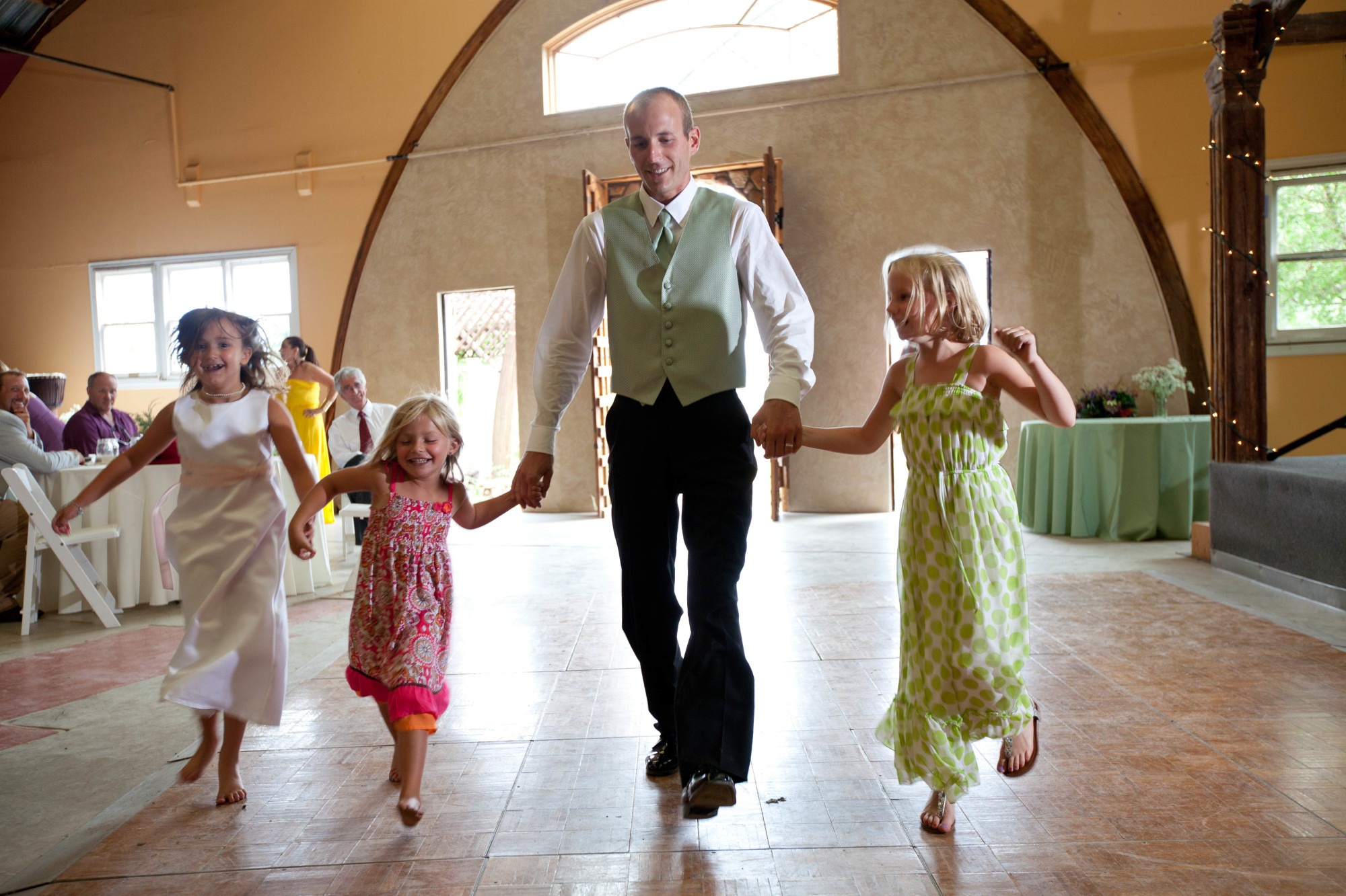 a groom in a green vest holds hands and skips with three girls