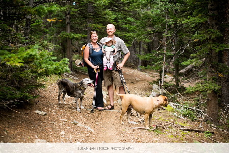 a mom, dad, daughter, and two dogs pose for a photo while hiking under pine trees