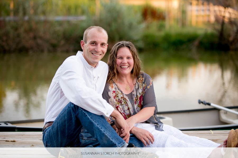 a smiling man and woman sit in evening light in front of a pond