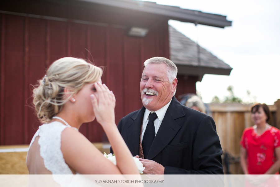 a man with facial hair laughs while a woman with blonde hair brushes it out of her eye in front of a red barn