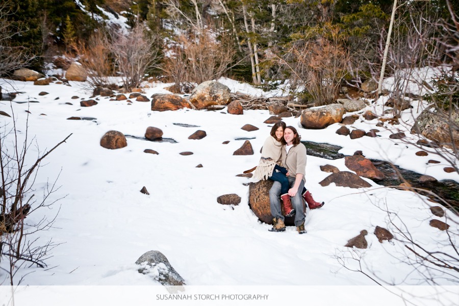 a couple sit on a big boulder on a snow-covered creekside