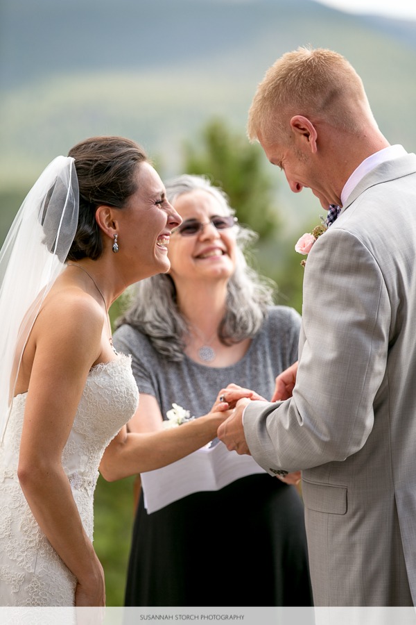 a groom slips a ring on a smiling brunette bride at an outdoor wedding ceremony in the mountains