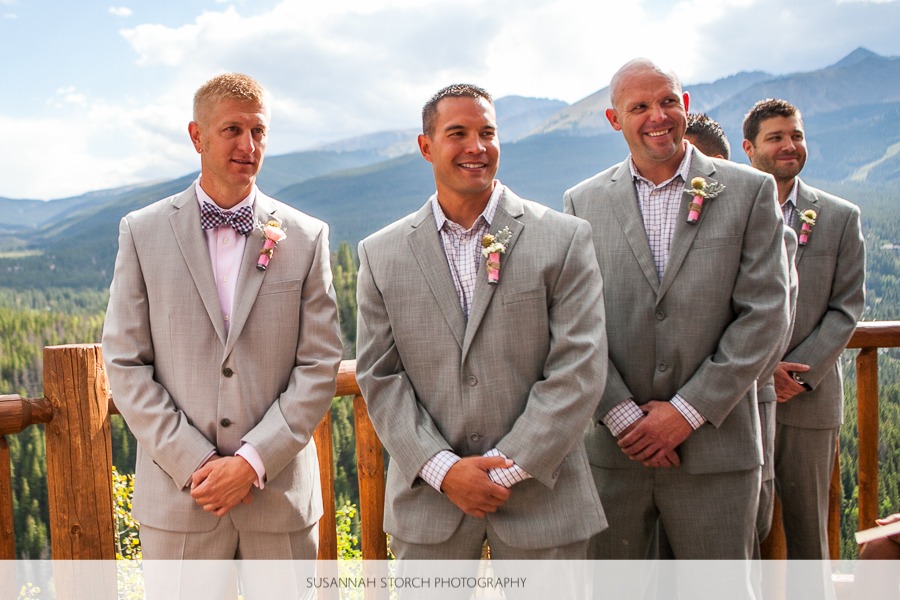 the groom and groomsmen stand on a deck in front of mountains and await the bride to walk down the aisle
