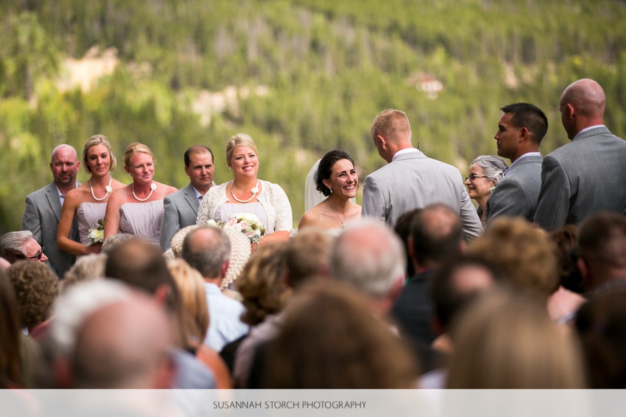 a bride smiles at her soon-to-be husband while her friends stand behind her at an outdoor mountain wedding ceremony