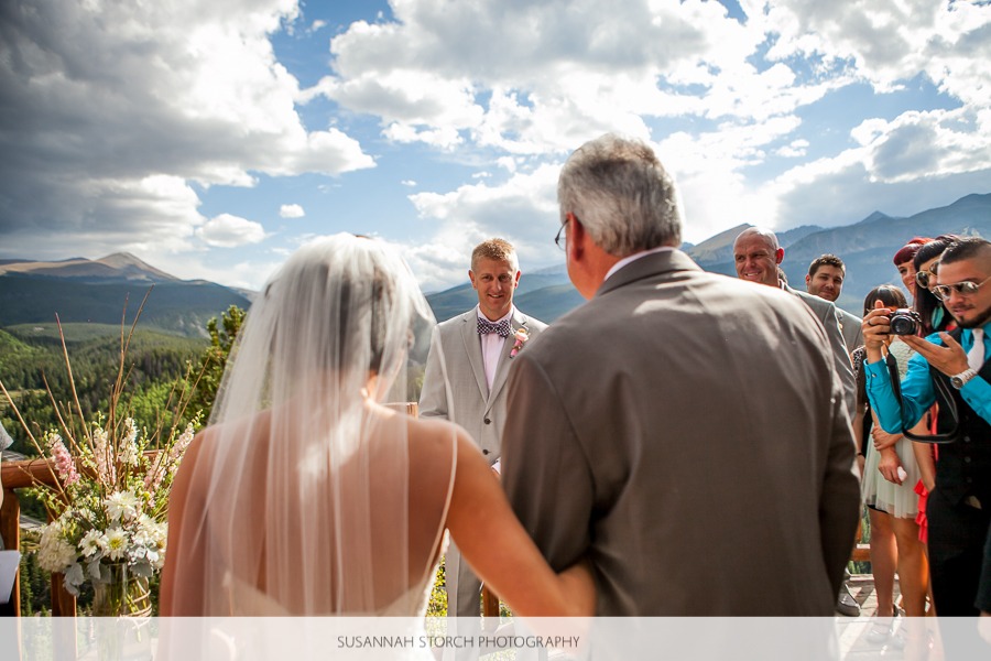 a bride and her dad walk down the aisle towards a blonde groom under dramatic clouds