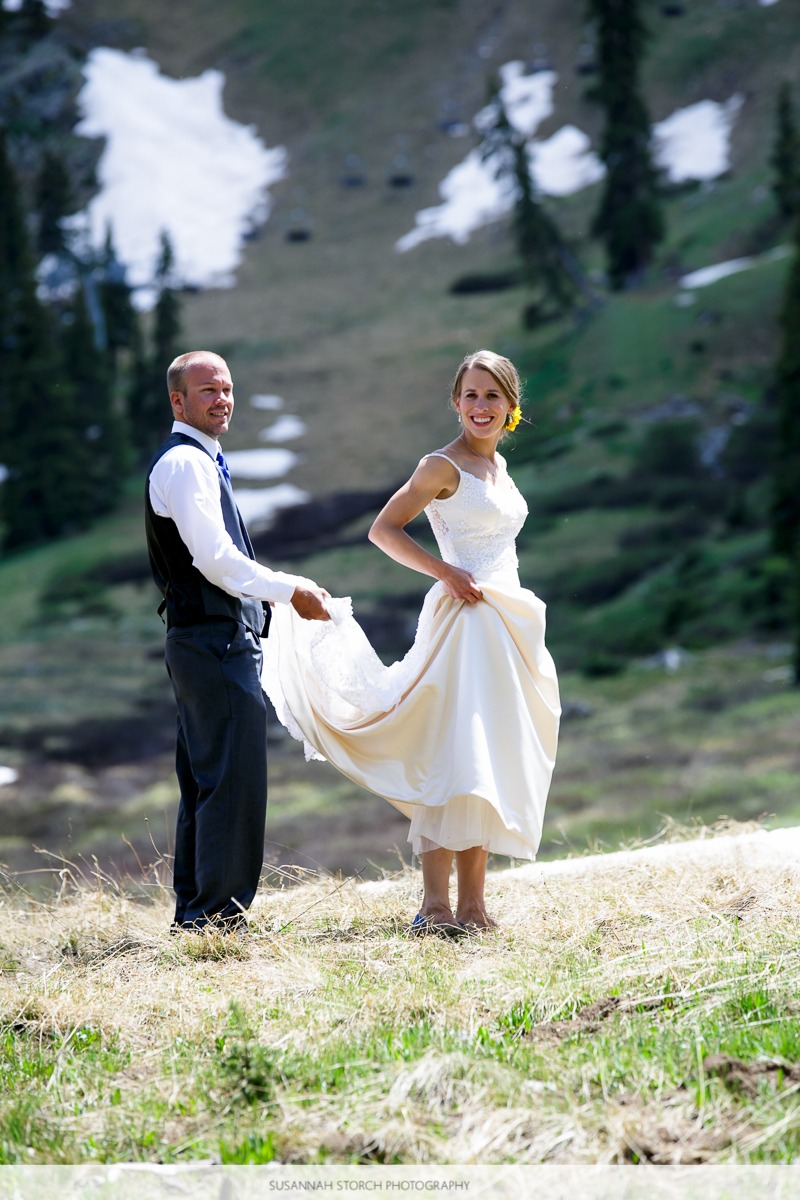 a groom lifts a bride's dress as they walk through grass