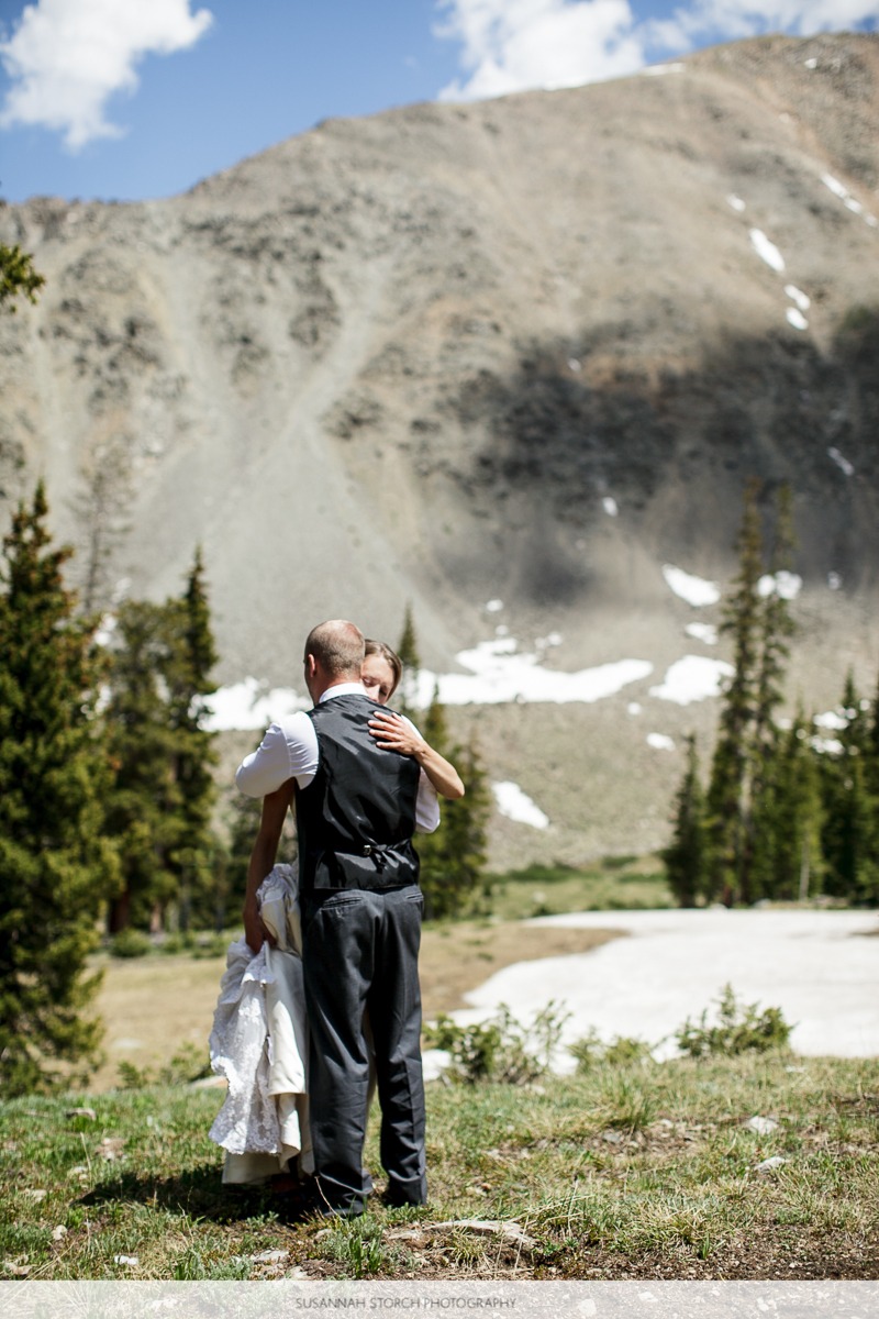a wedding couple hug while standing in front of pine trees, a snow field, and rugged mountains