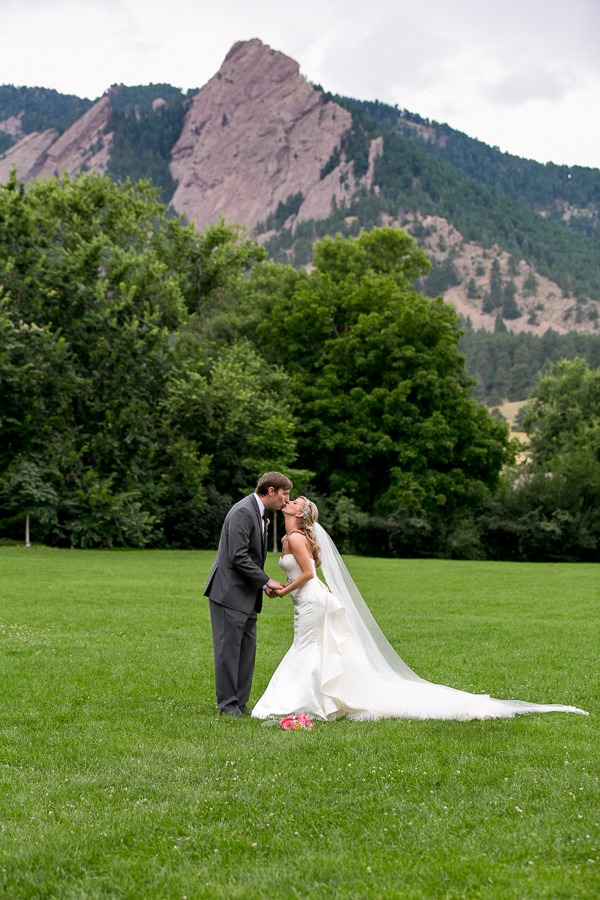 a bride and groom kiss on a green lawn in front of boulder's flatirons