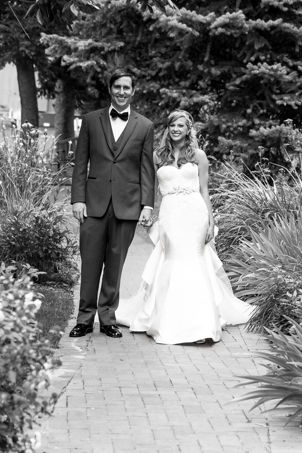 black and white photo of a groom in a bowtie and a blonde bride walking down a brick path
