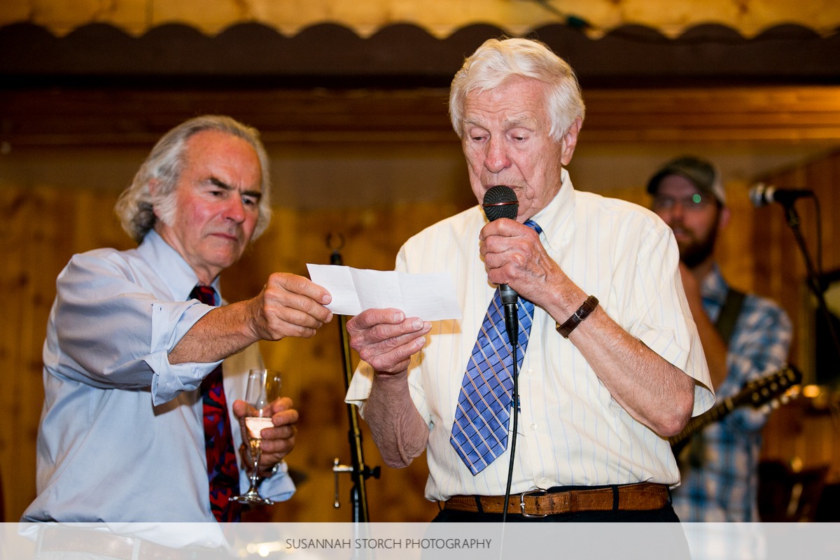 an older gentleman holds onto a piece of paper with the help of another man and reads a speech while holding a microphone
