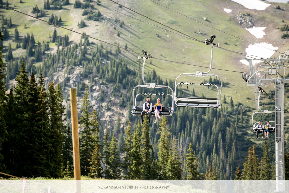 a man and woman ride on a ski lift in front of rugged mountains