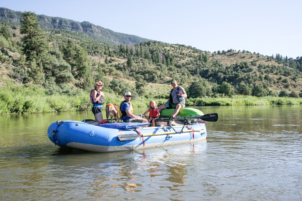 four people sitting on a blue raft on calm water in front of arid mountains