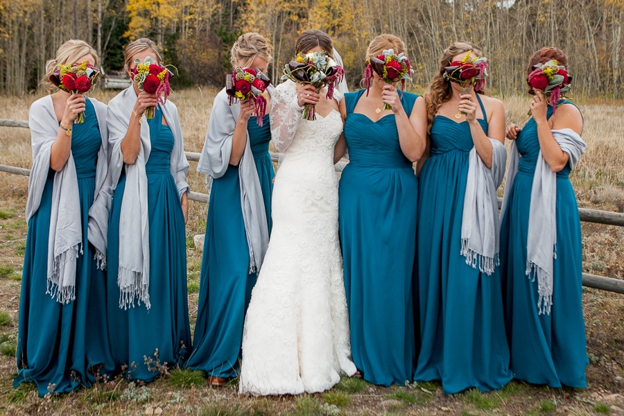 a line of bridesmaids in blue dresses surround the bride and all hold red bouquets in front of their heads