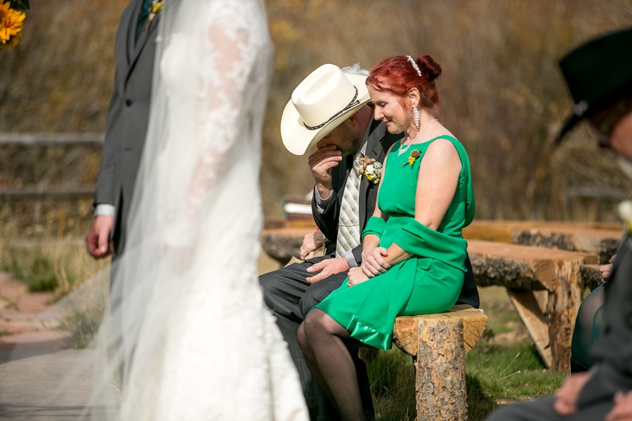 a woman with red hair and a man in a hat sit on a log bench during an outdoor wedding ceremony