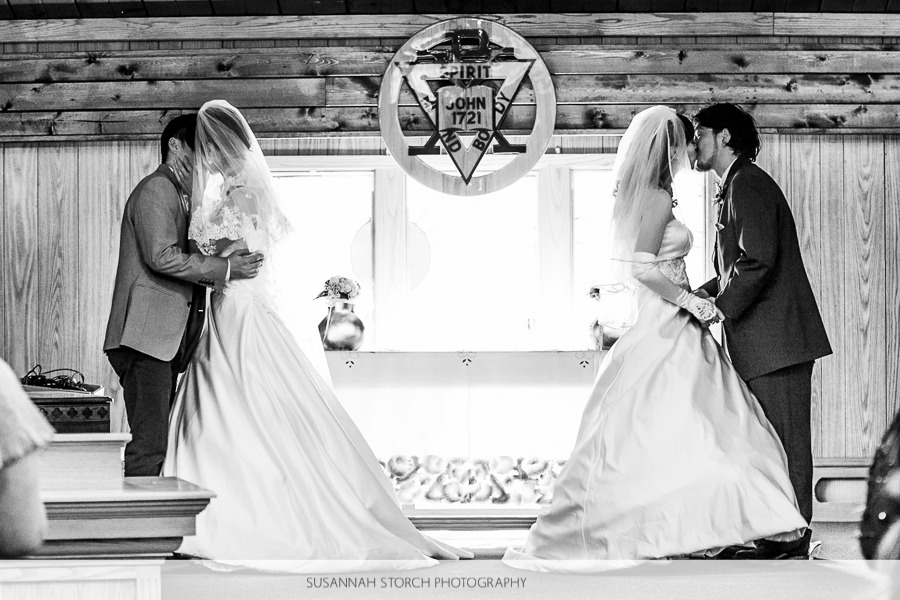 black and white photo of two wedding couples kissing in front of a window