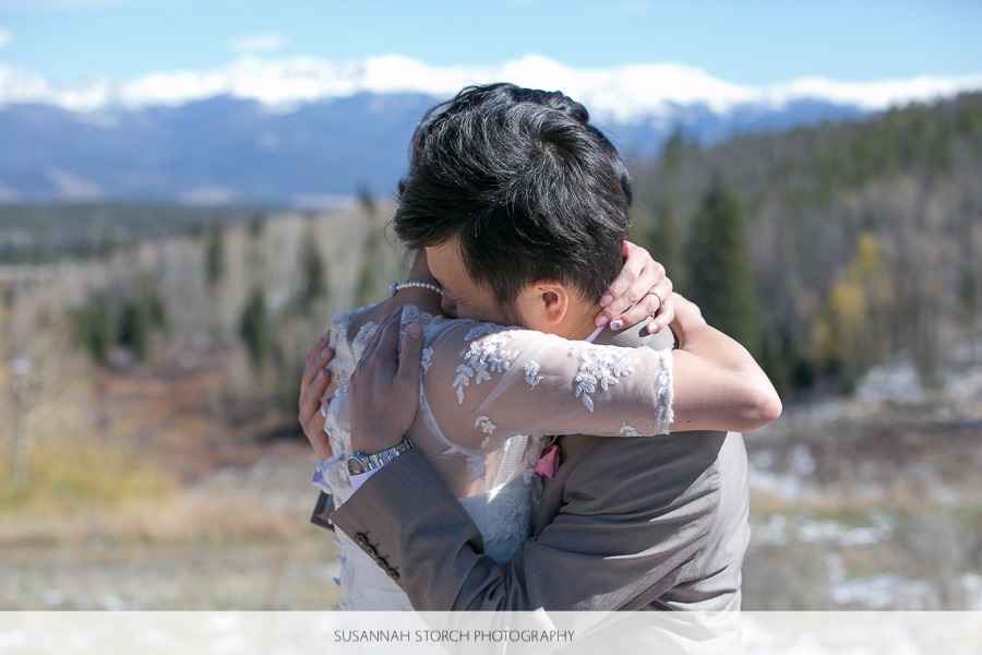 a bride in a lacy shawl hugs her groom in front of snow-capped mountains