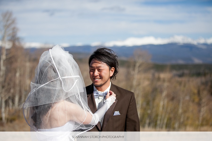 veiled bride adjusts the bowtie of her groom while standing outside near snow-capped mountains