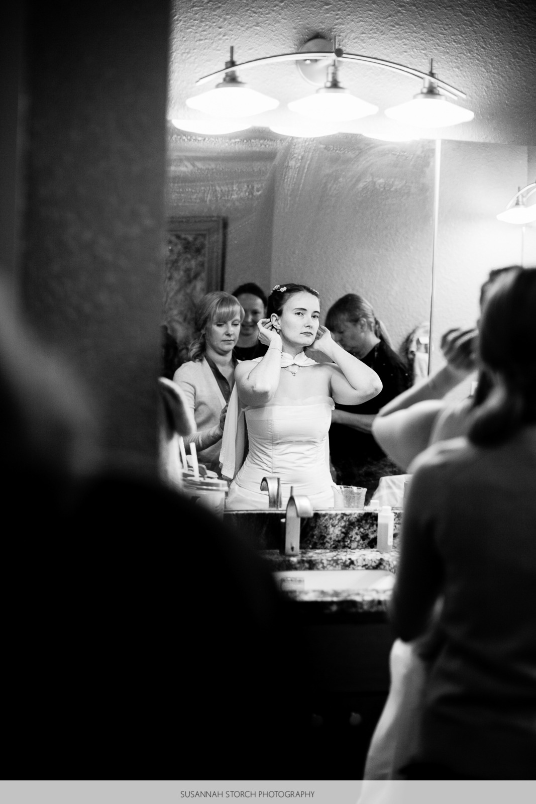 black and white photo of a bride looking into a mirror as she gets ready with the help of other women