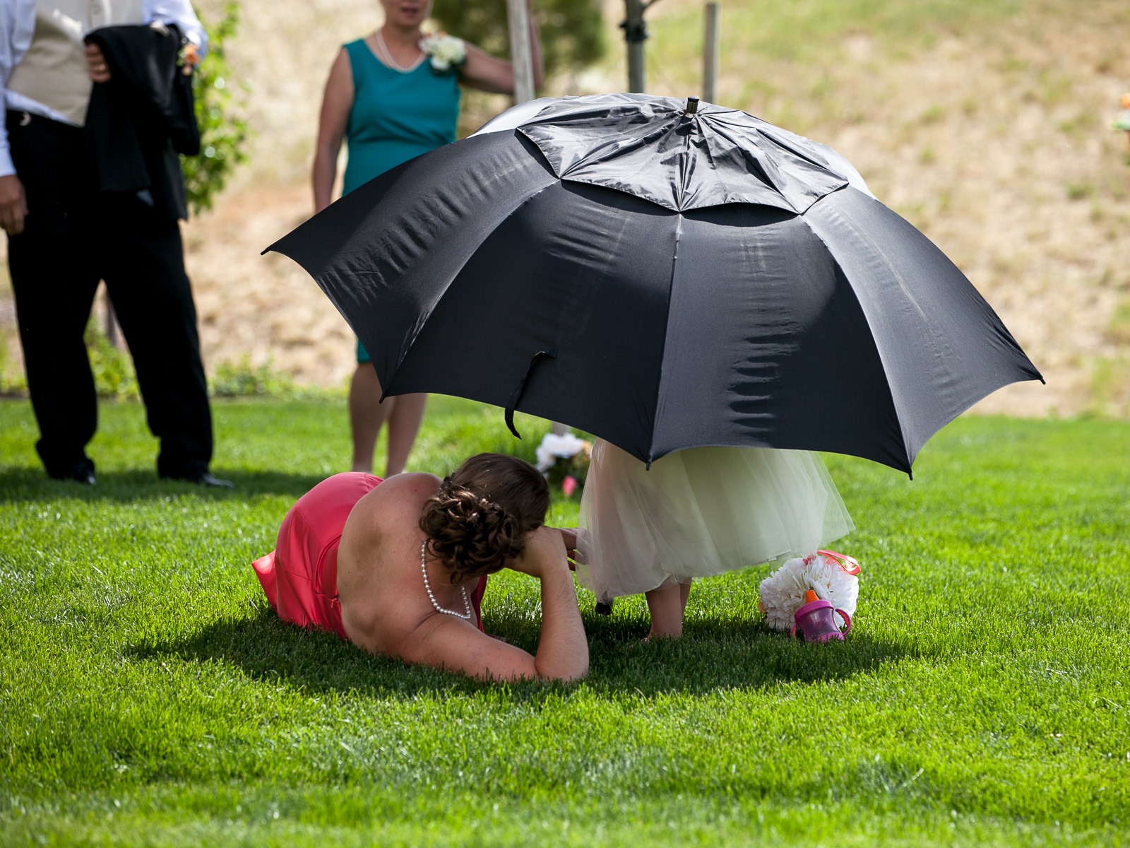 a flower girl stands under a black umbrella on a green lawn to escape the sun