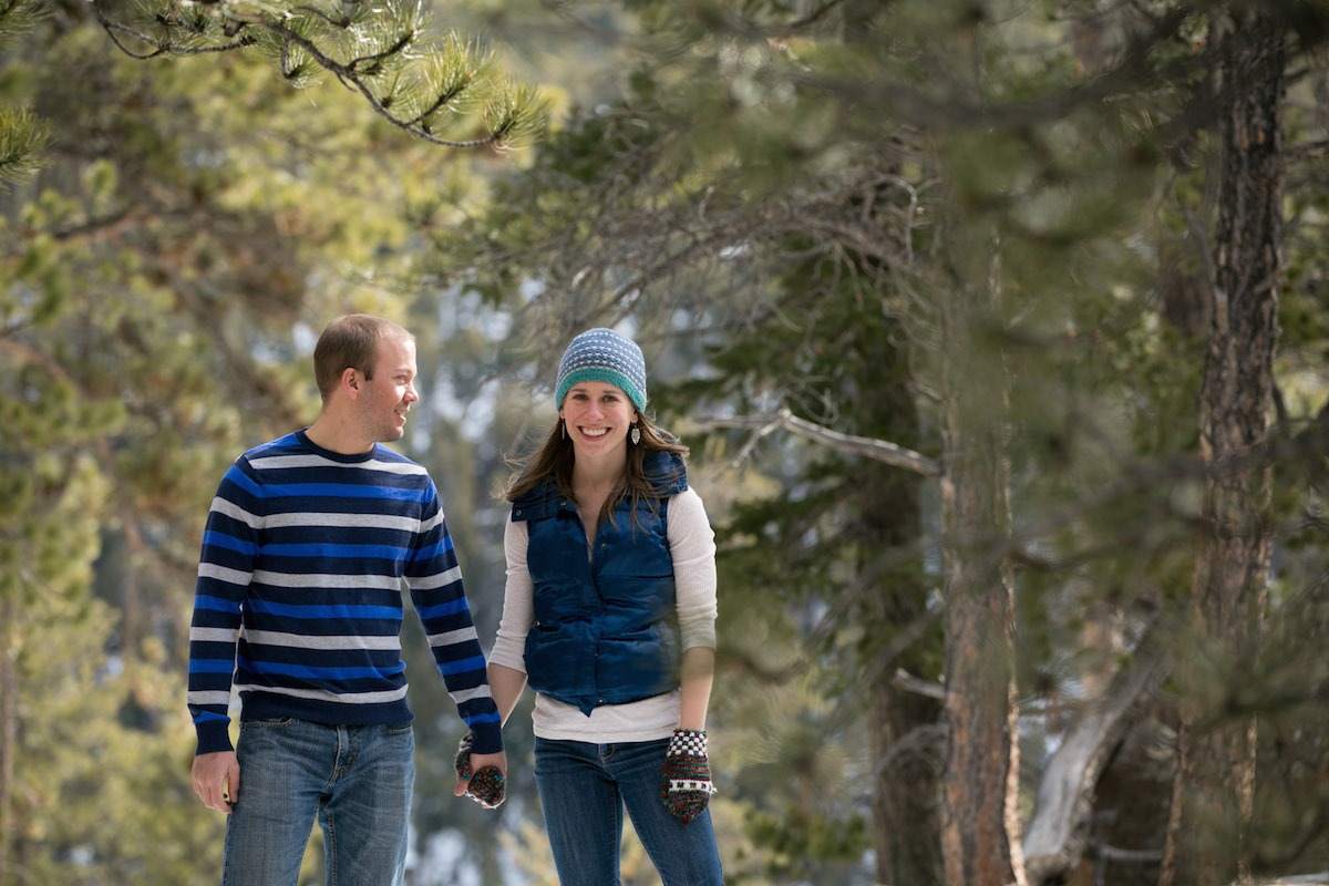 a man looks at a woman wearing a knit hat and gloves while standing among pine trees