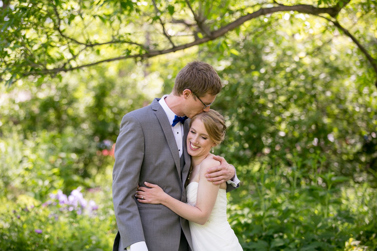 a tall man in a gray suit kisses the top of a bride's head in front of green gardens and trees