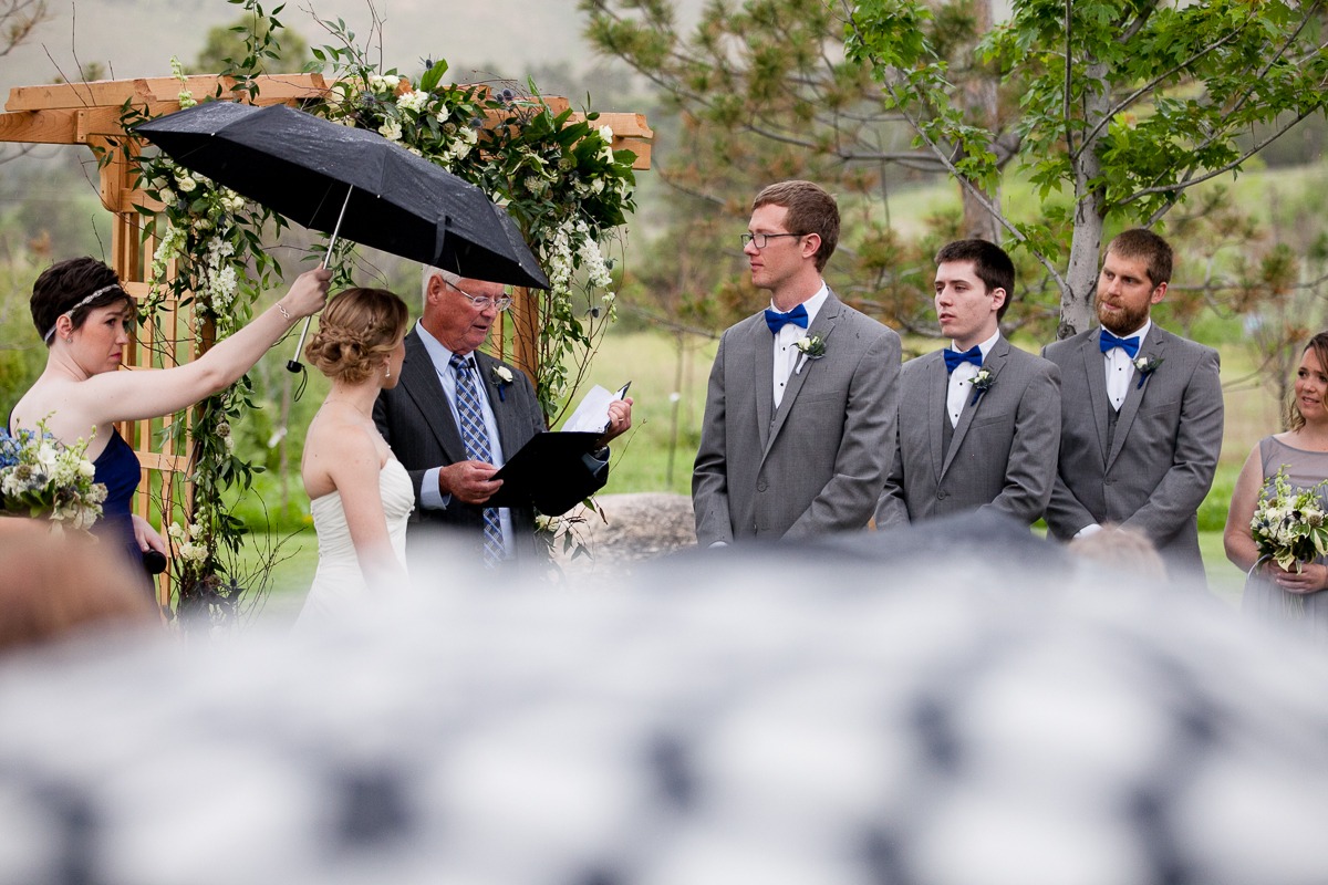 a woman holds an umbrella over a bride during an outdoor wedding ceremony