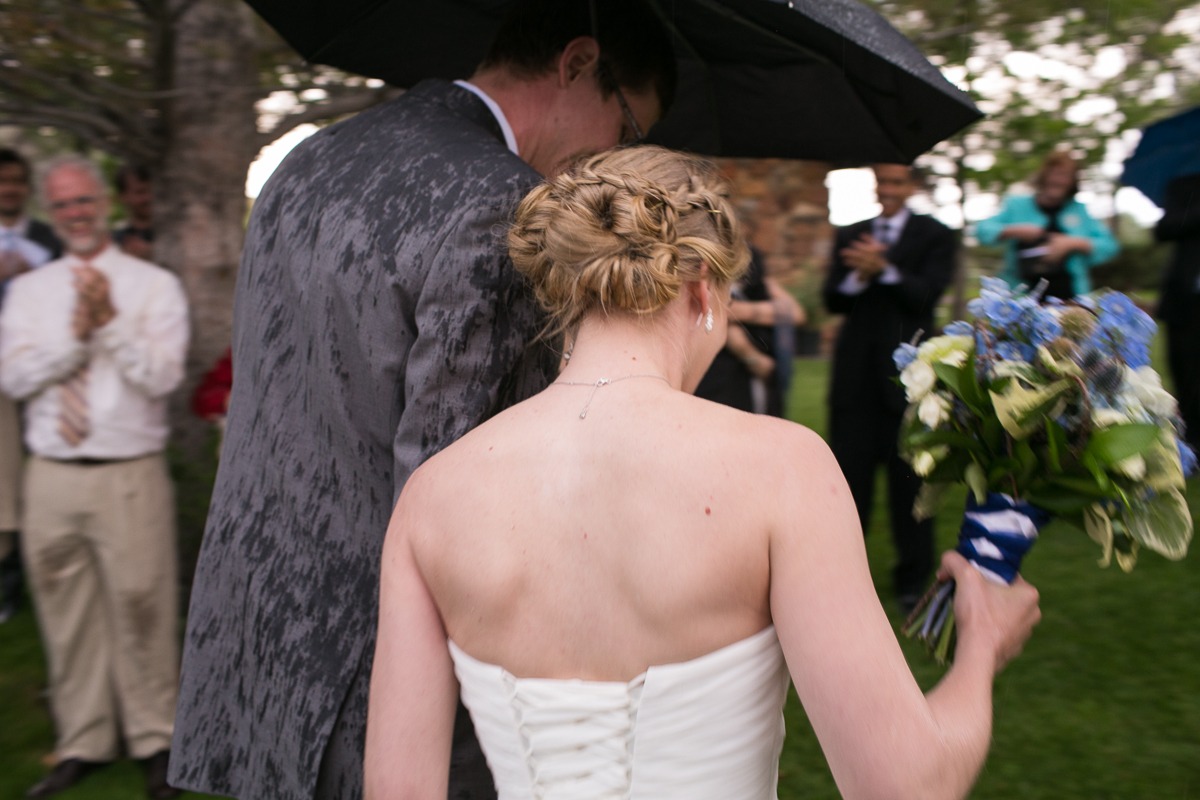 a groom carries an umbrella and a bride holds flowers as they walk away from the camera in the rain