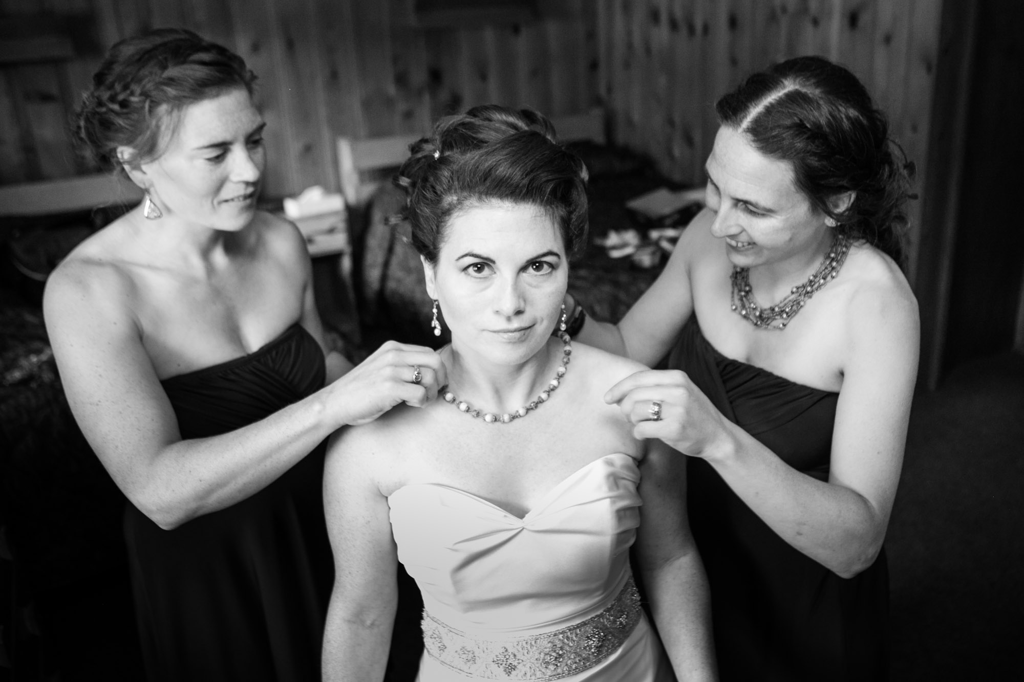a bride looks at the camera while her bridesmaids stand behind her and fuss with a necklace