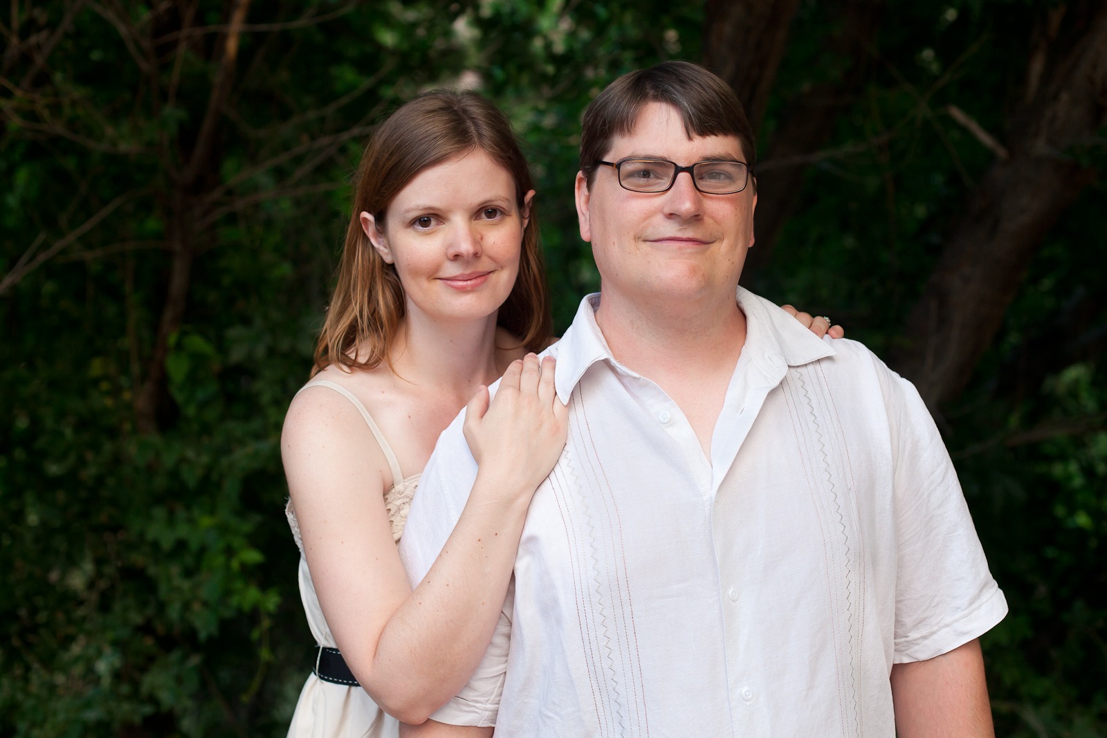 an engaged couple in white and cream stand in front of dark trees