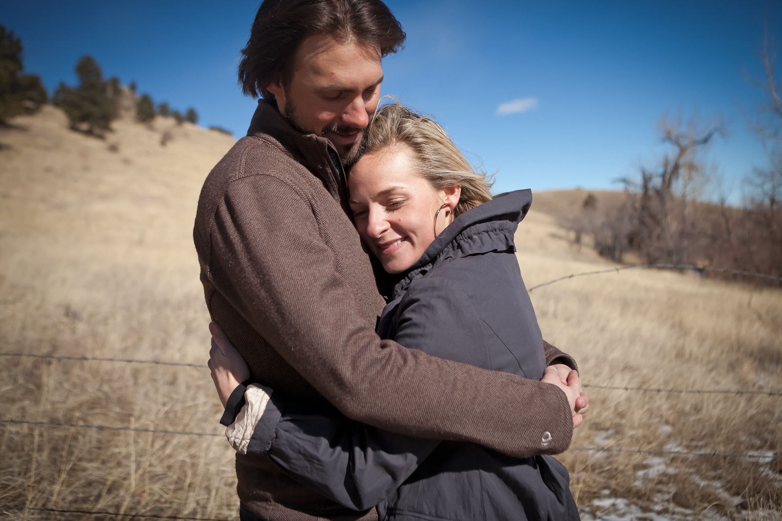 A couple wearing brown hug in front of dried brown grass under very blye skies