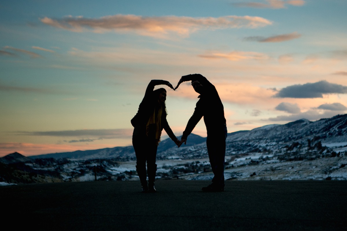 silhouette of a heart made out of people's hands in front of snowy mountains at dusk