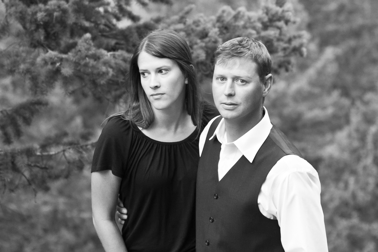 a black and white image of an engaged couple looking serious while standing in front of pine branches