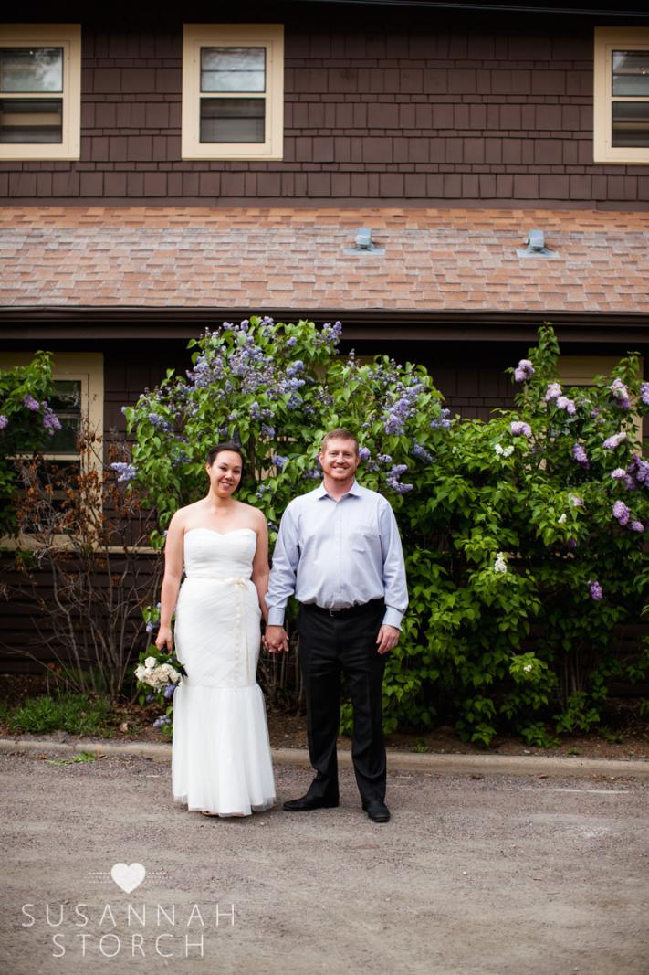 a bride and groom hold hands in front of a lilac bush