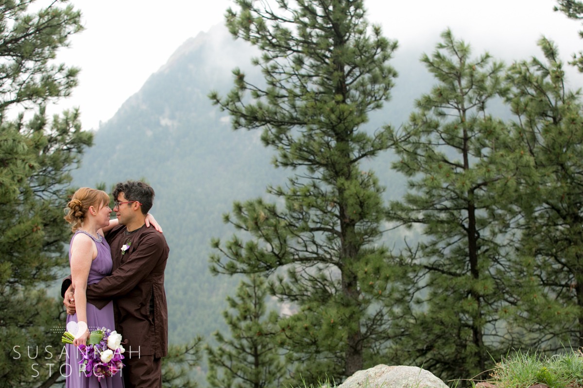 a bride and groom face each other in front of green, foggy mountains