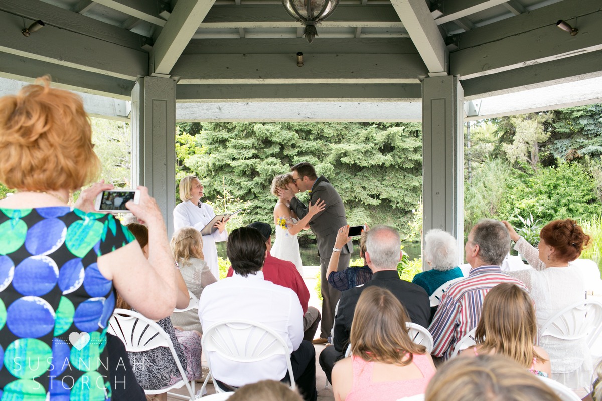 a bride and groom kiss as wedding guests watch and capture the moment with their digital cameras