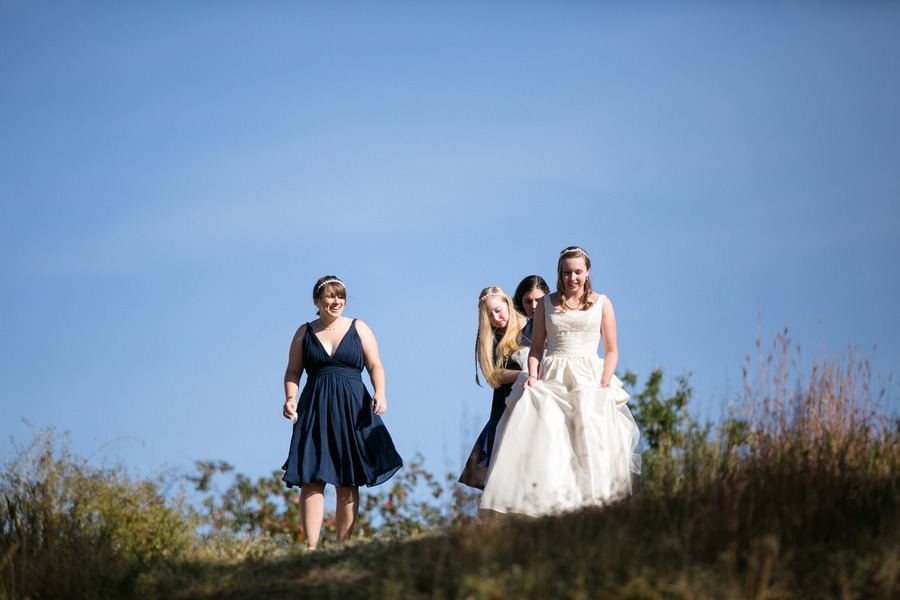 a bride walks fown a hill in front of blue skies with the help of two ladies