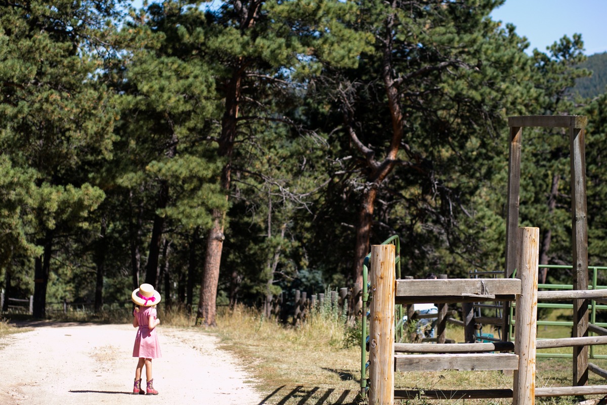 a young girl in cowboy boots and a hat stands on a dirt road