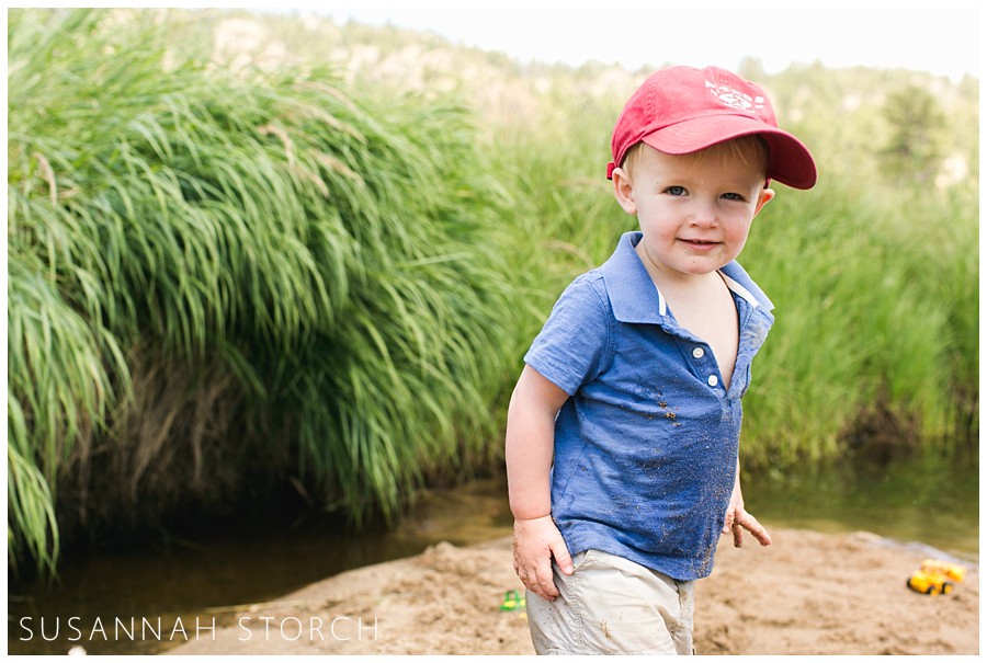 a toddler in a red hat smiles while standing on a stream bank