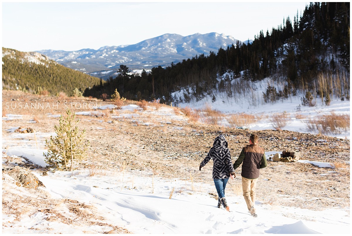 a couple hold hands and walk away from the camera in a mountain landscape