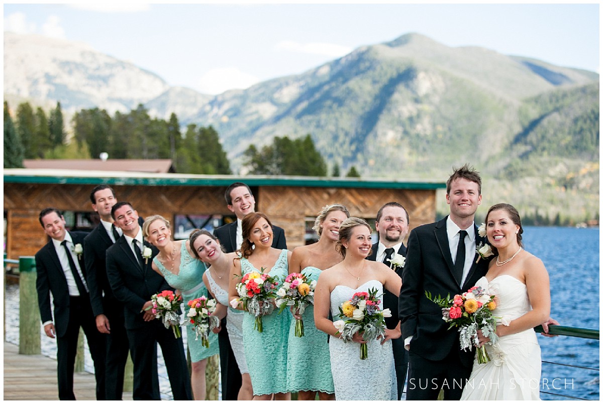 A bride and groom stand by a mountain lake as the bridal party members look at them