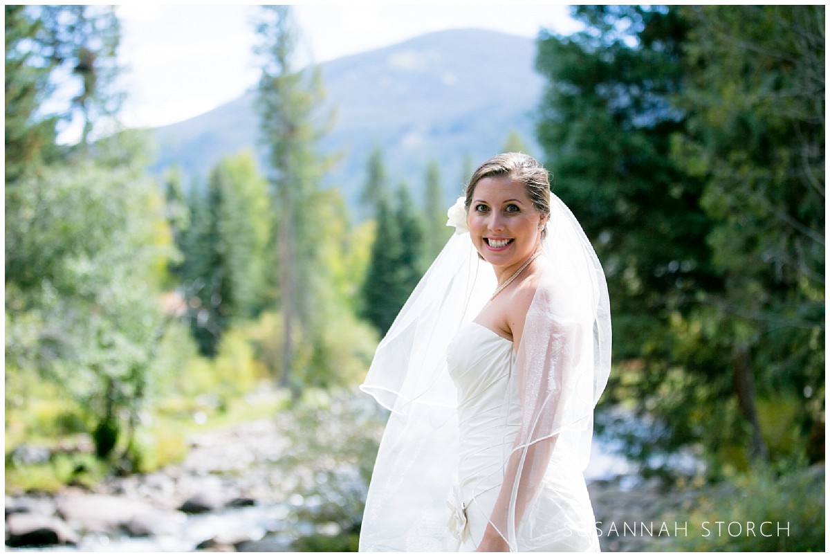 a bride poses in front of a stream, pine trees, and mountains